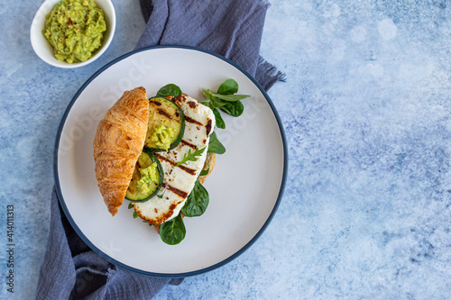 Croissant sandwich with grilled halloumi cheese, arugula and grilled zucchini, blue concrete background. Healthy breakfast or lunch. photo