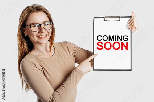 COMING SOON. Beautiful young business woman wearing glasses holds a clipboard with mock up space isolated on gray background