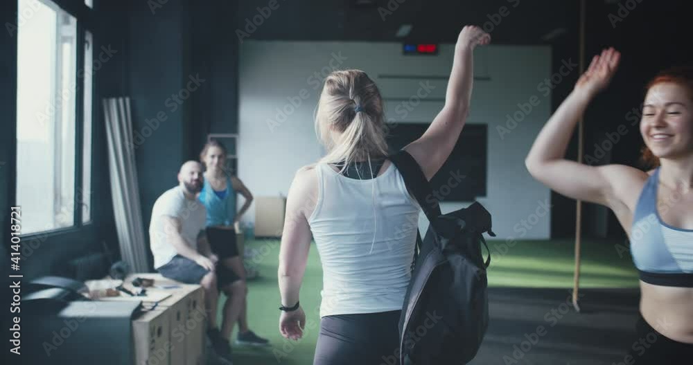 Lifestyle fitness community club. Camera follows young sporty blonde woman enter modern gym, high-fiving happy friends.