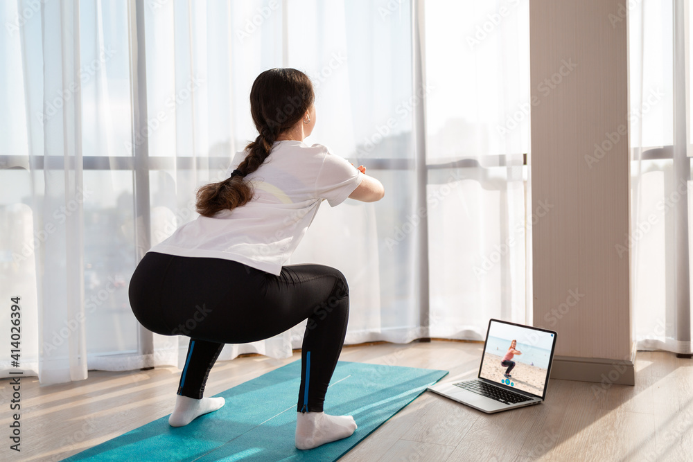 A young woman in sportswear trains at home for online lessons with trainer. Rear view. Indoor. Concept of home online training. A young woman in sportswear trains at home for online lessons with trainer. Rear view. Indoor. Concept of home online training.