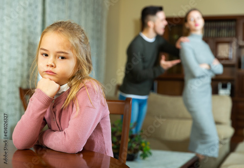 Upset little girl sitting alone while her young parents quarreling at living room