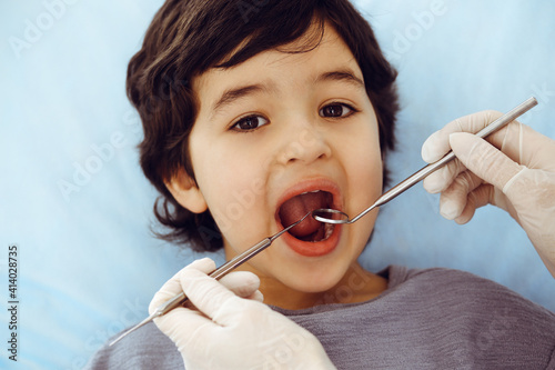 Cute arab boy sitting at dental chair with open mouth during oral checking up with doctor. Visiting dentist office. Stomatology concept