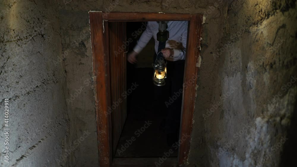 Covid-19, post-apocalypse. A man in a gas mask, white shirt, tie, and kerosene lamp opens a wooden door in a concrete bunker and waves to enter.