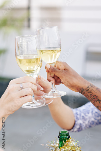 Close-up image of women clinking glasses of white wine over bottle wrapped in tinsel when celebrating Christmas