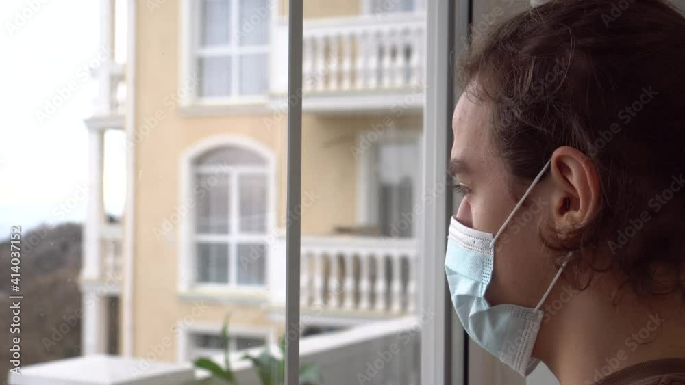 Self care self isolation quarantine lockdown during Covid-19. Alone sad young man in a mask looks out the window