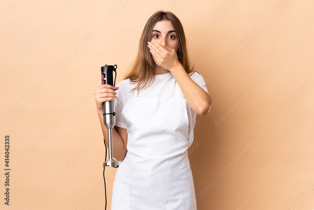 Woman using hand blender over isolated background covering mouth with hands Woman using hand blender over isolated background covering mouth with hands
