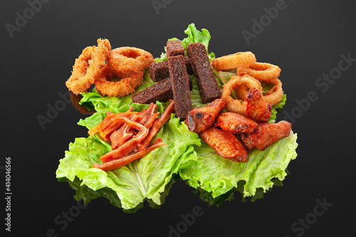 A set of snacks for beer. Grilled chicken wings, onion rings, squid rings, garlic croutons, dried pork ears. On a lettuce leaf. Decorated with greenery. Close-up. Dark mirror background. photo