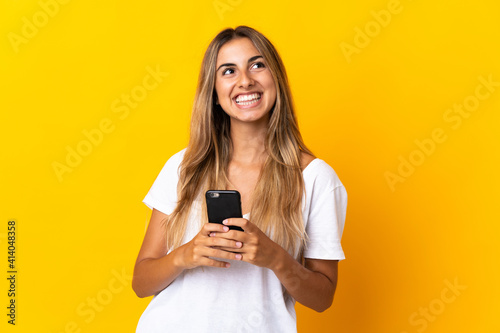 Young hispanic woman over isolated yellow background using mobile phone and looking up