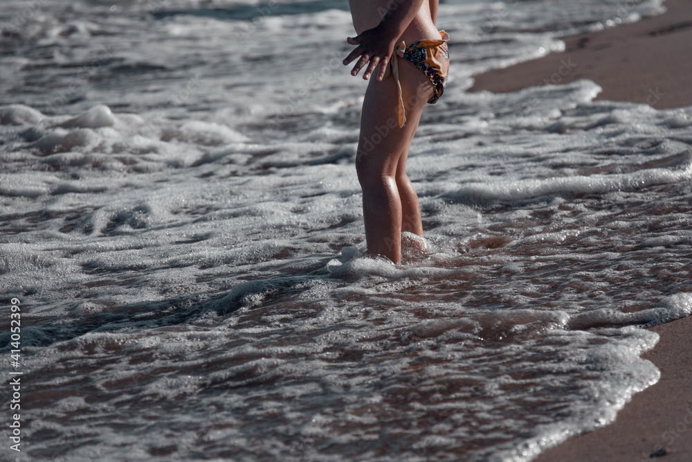Fototapeta premium A child on the beach. Children's feet on the background of the contrasting sun are immersed in the sand, which is washed by a wave of sea foam. Family vacation.