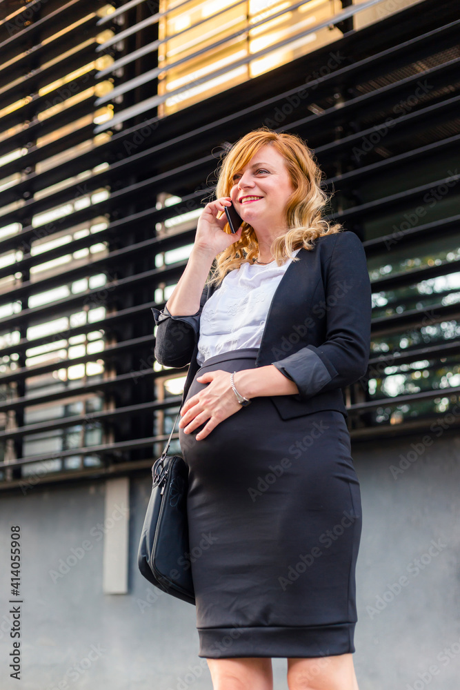 Fototapeta premium A smiling pregnant businesswoman standing in front of a modern building having a phone call.
