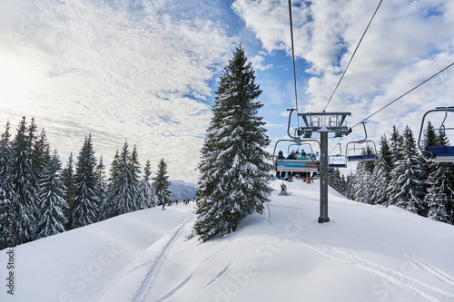 Panorama of snowy hills with coniferous trees, ski lifts and people under blue cloudy sky. Scenery of snow-covered slopes with chairlift mechanism for transporting skiers. Concept of ski resort.