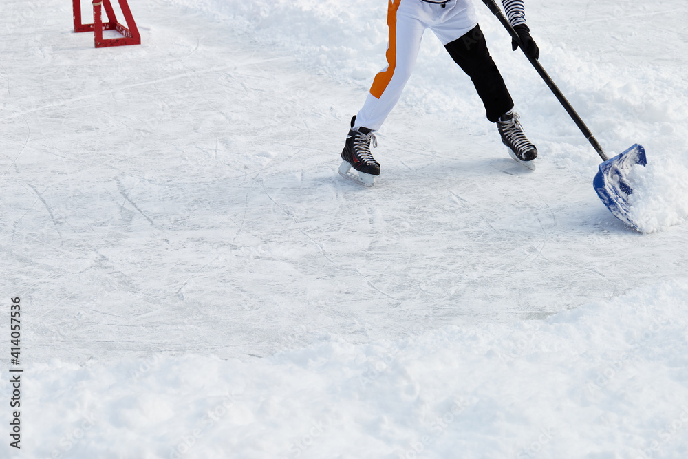 Hockey player on ice-skating removing snow with a blue shovel in the winter in a park. Wearing white clothes. Moving from goal to corner. Ice rink cleaning. Active sport outdoor. Hockey player on ice-skating removing snow with a blue shovel in the winter in a park. Wearing white clothes. Moving from goal to corner. Ice rink cleaning. Active sport outdoor.