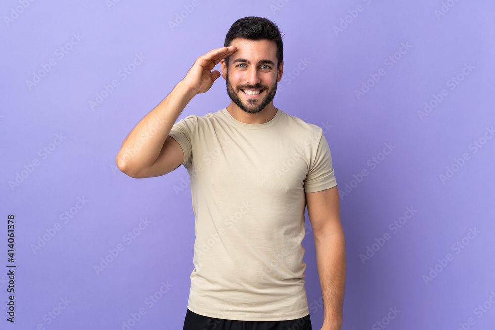 Young handsome man with beard over isolated background saluting with hand with happy expression Young handsome man with beard over isolated background saluting with hand with happy expression