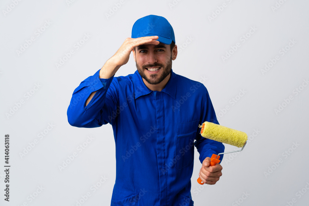 Painter man holding a paint roller isolated on white background looking far away with hand to look something