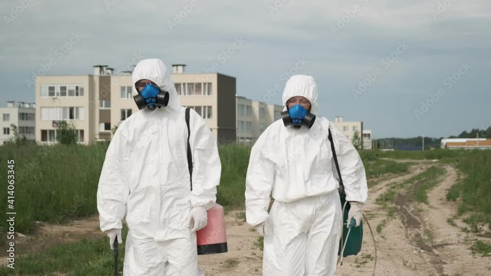 Two people in respirators and protective overalls walk along road into field.