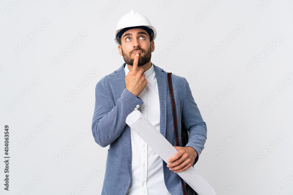 Young architect man with helmet and holding blueprints isolated on white background having doubts while looking up Young architect man with helmet and holding blueprints isolated on white background having doubts while looking up