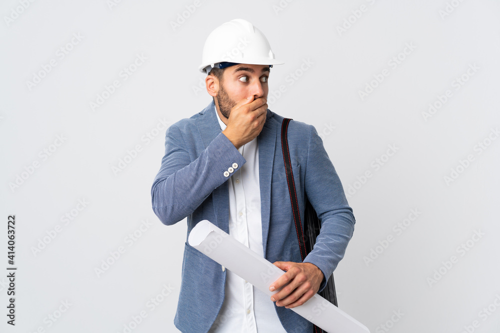 Young architect man with helmet and holding blueprints isolated on white background covering mouth and looking to the side Young architect man with helmet and holding blueprints isolated on white background covering mouth and looking to the side