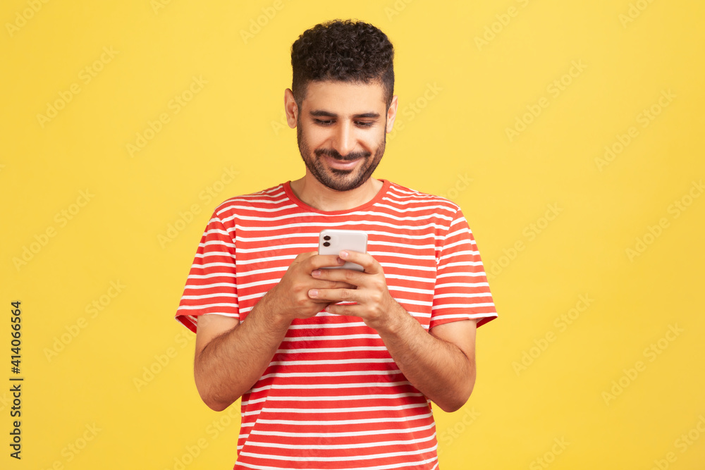 Smiling happy bearded man in striped t-shirt texting on smartphone, chatting in social networks or having online conversation in messenger. Indoor studio shot isolated on yellow background Smiling happy bearded man in striped t-shirt texting on smartphone, chatting in social networks or having online conversation in messenger. Indoor studio shot isolated on yellow background
