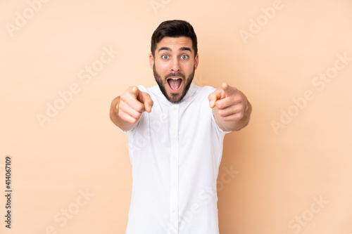 Caucasian handsome man isolated on beige background surprised and pointing front
