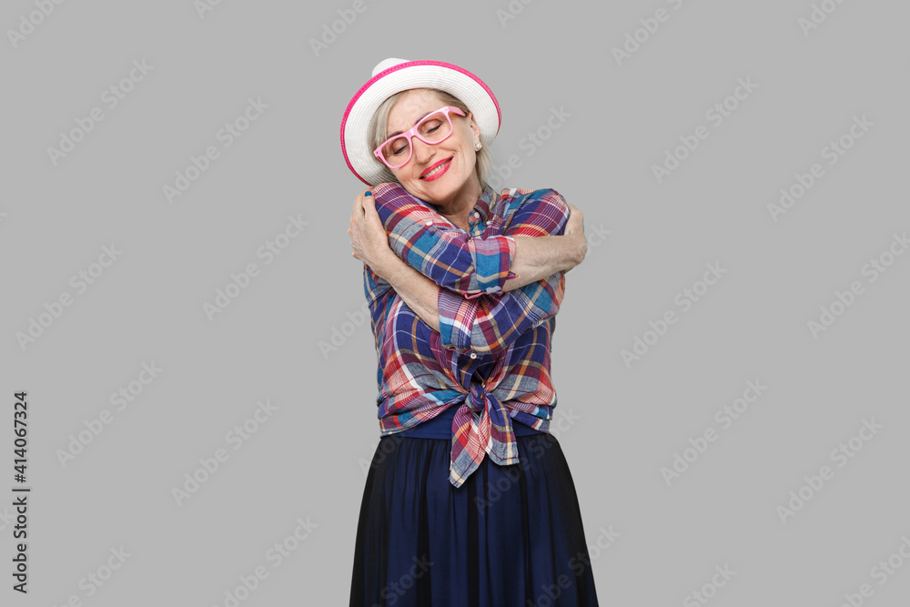 Portrait of peacful happy modern stylish mature woman in casual style with hat, eyeglasses standing, hugging herself with closed eyes and toothy smile. indoor studio shot isolated on gray background