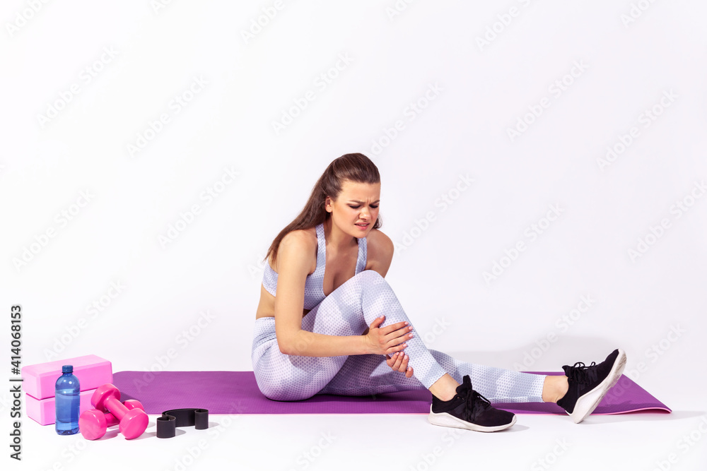 Full length athletic woman in white sportswear sitting on gym mat touching injured caviar, suffering calf muscle strain after yoga workout. Indoor studio shot isolated on gray background Full length athletic woman in white sportswear sitting on gym mat touching injured caviar, suffering calf muscle strain after yoga workout. Indoor studio shot isolated on gray background