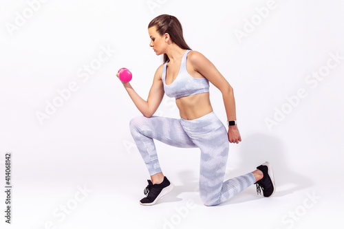 Full length side view athletic woman in white sportswear holding dumbbells in hand doing squats with one leg, standing on knee. Indoor studio shot isolated on gray background