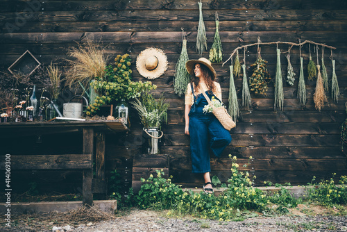 Attractive girl wearing straw hat and blue denim dungarees relaxing near wooden old summerhouse wall on sunny day.