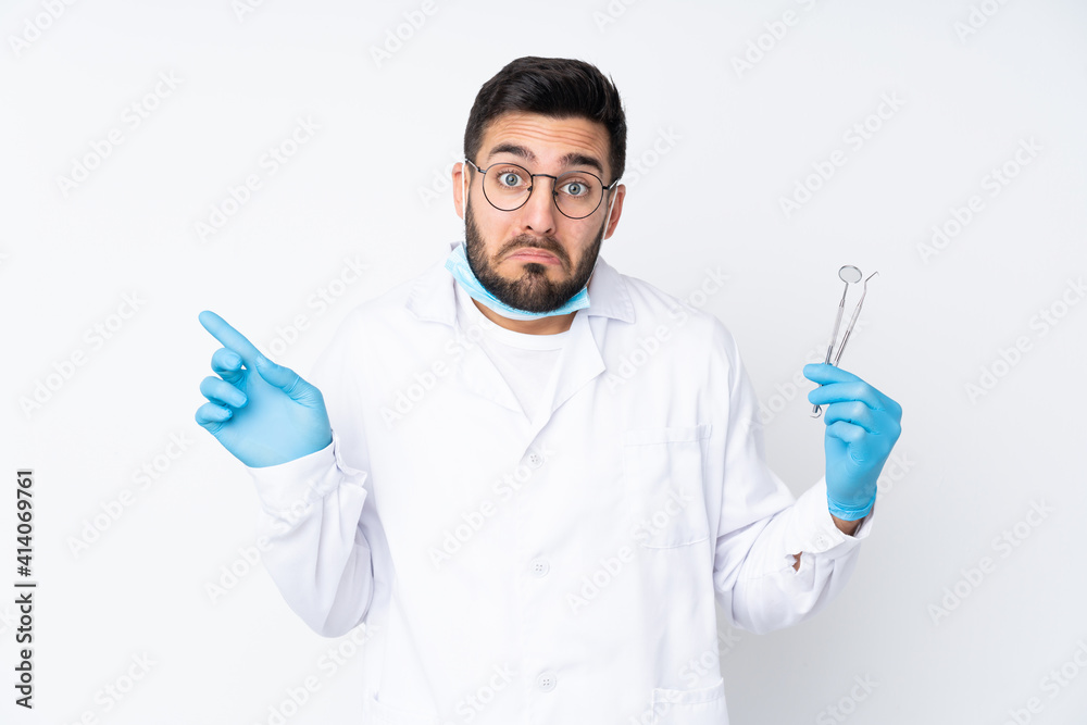 Dentist man holding tools isolated on white background pointing to the laterals having doubts