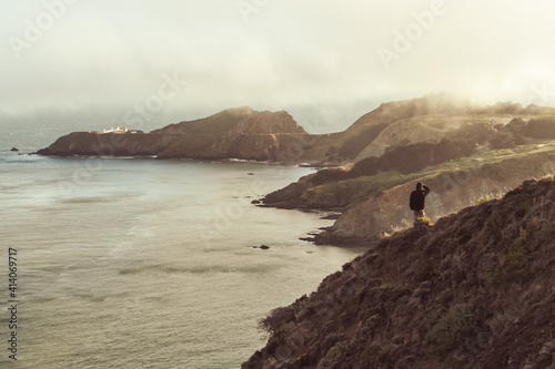 Man in outdoor cloths standing alone on a hill by the ocean overlooking the Point Bonita Lighthouse on a foggy San Francisco day, California, United States of America, USA