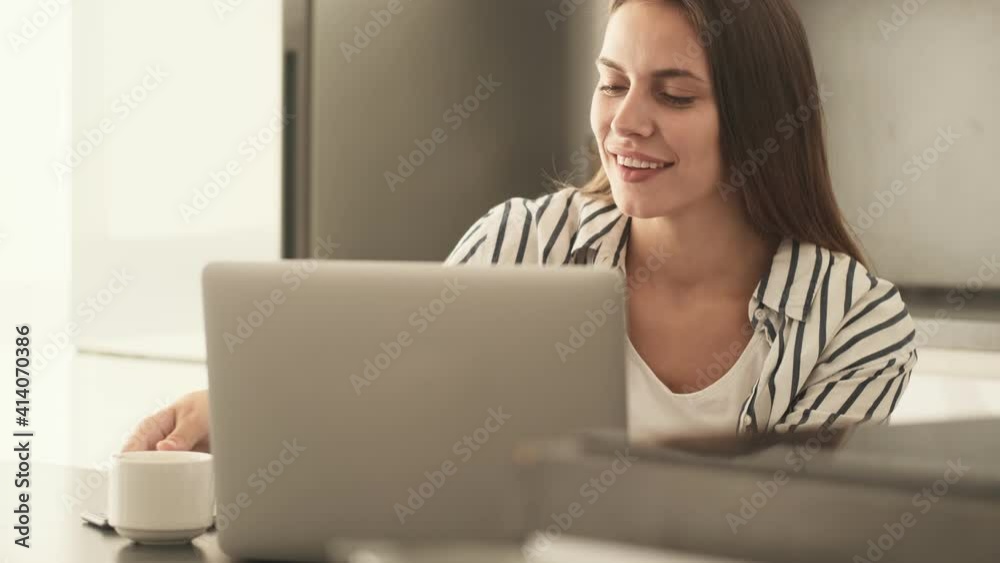 A smiling young woman is using her laptop computer while sitting in the kitchen at home A smiling young woman is using her laptop computer while sitting in the kitchen at home