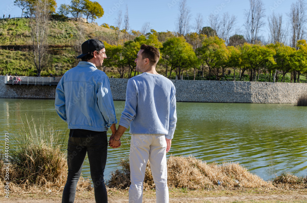 Romantic Scene of Casual Gay Couple Lovers Looking at Each Other and Holding Hands in front of a Lake at the Park Romantic Scene of Casual Gay Couple Lovers Looking at Each Other and Holding Hands in front of a Lake at the Park