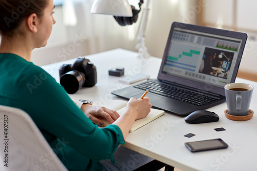 technology, post production and vlog concept - close up of woman working in video editor program on laptop computer writing to notebook at home office