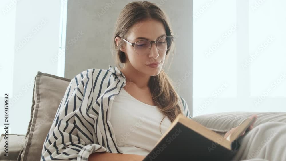 A charming young woman wearing glasses is reading book sitting on the sofa at home A charming young woman wearing glasses is reading book sitting on the sofa at home