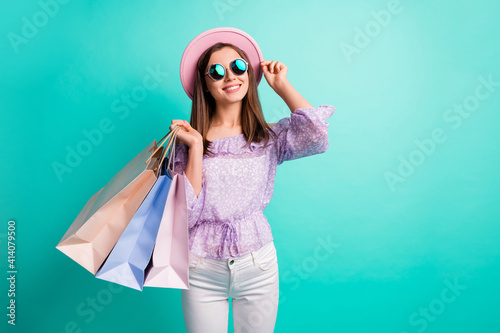 Photo of optimistic brunette girl hold bags look empty space hand cap wear purple shirt trousers spectacles isolated on blue color background
