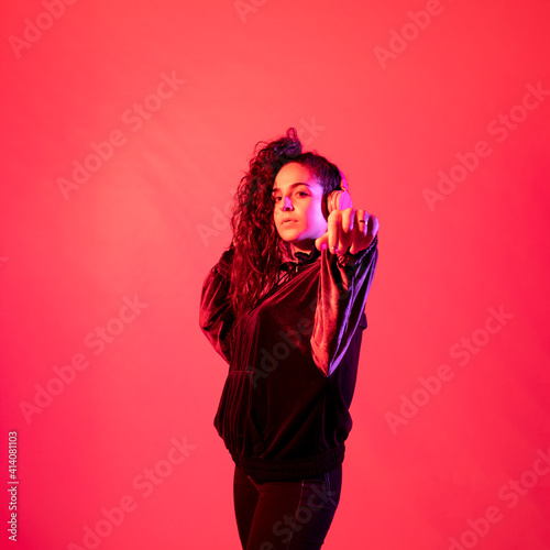 Vertical shot of a young girl with her brown curly hair in a ponytail wearing headphones pointing her hand to the camera, against red background © Noelia