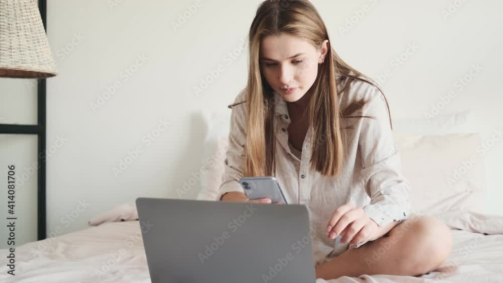A focused woman is using her laptop computer and her smartphone while sitting in the bed at the white bedroom at home A focused woman is using her laptop computer and her smartphone while sitting in the bed at the white bedroom at home