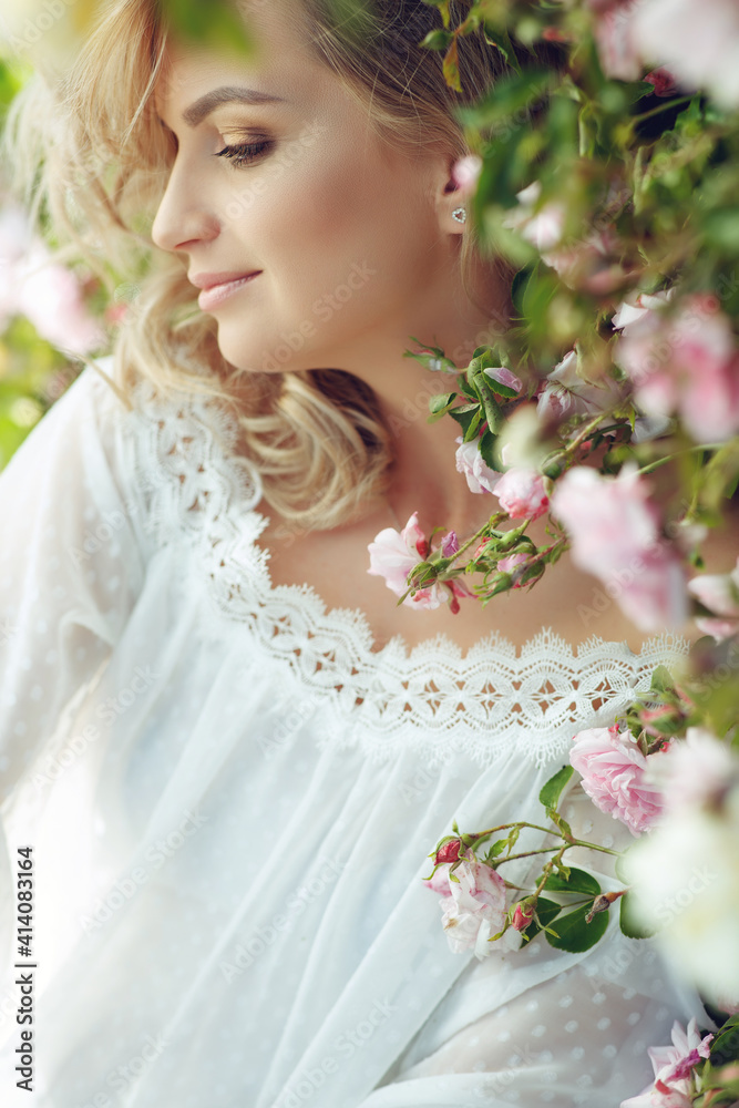 Fototapeta premium Beautiful young woman with long curly hair and perfect skin posing near blooming roses in the garden. Nude makeup. Close up portrait. High quality photo.