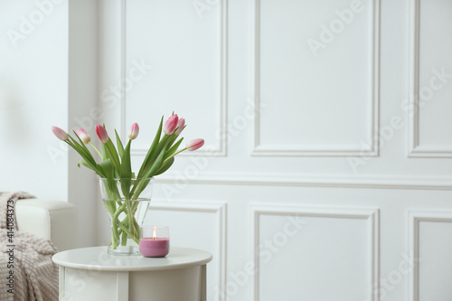 Beautiful tulips and burning candle on white table indoors  space for text. Interior design