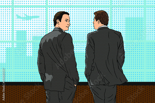 Two young business men in suits stand in the office near the window, look at the city and have a conversation.