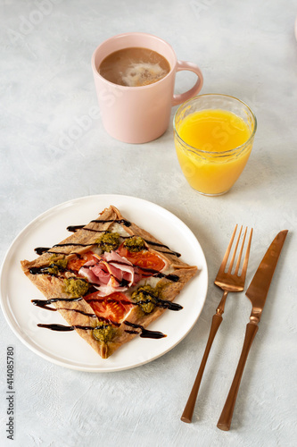 Galette bretonne with emmental cheese, homemade pesto, tomatoes and bacon. A mug of coffee and a glass of orange juice on the table.