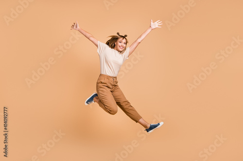 Full length photo of girl jump having fun wear t-shirt pants sneakers isolated beige color background