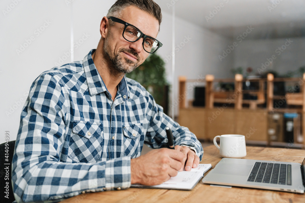Pleased grey man writing down notes while working with laptop Pleased grey man writing down notes while working with laptop