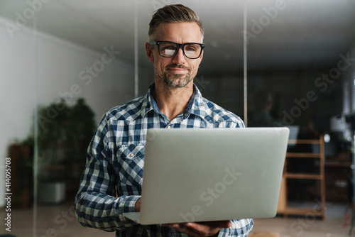 Pleased handsome man working with laptop while standing in office