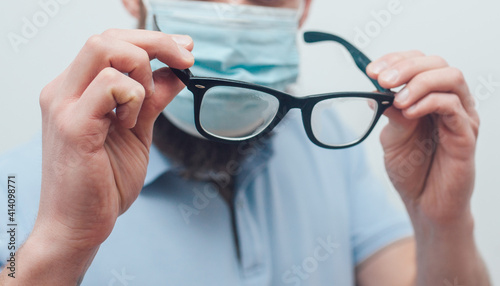 Handsome man with beard wiping blurred foggy misted glasses caused by wearing medical mask on light background. Covid-19