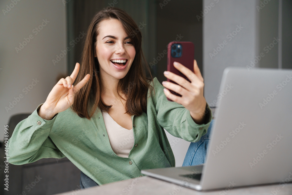 Cheerful woman showing peace sign while using cellphone and laptop Cheerful woman showing peace sign while using cellphone and laptop