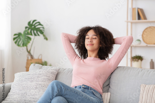 In cozy living room happy african american woman put hands behind head sitting leaned on couch