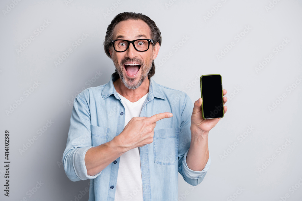 Portrait of handsome amazed cheerful mature man holding in hand demonstrating device isolated over grey pastel color background Portrait of handsome amazed cheerful mature man holding in hand demonstrating device isolated over grey pastel color background