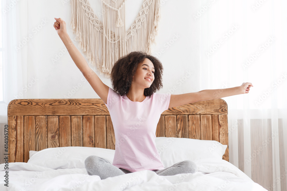 Happy teen african american fresh vitality lady sits on bed, stretching arms after sleep and enjoying morning Happy teen african american fresh vitality lady sits on bed, stretching arms after sleep and enjoying morning