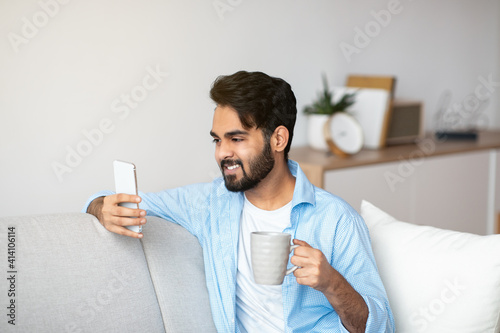 Domestic Leisure. Handsome Eastern Guy Using Smartphone And Drinking Coffee At Home