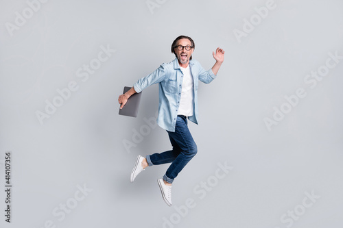 Full length body size view of handsome cheerful amazed man jumping carrying laptop running isolated over grey pastel color background